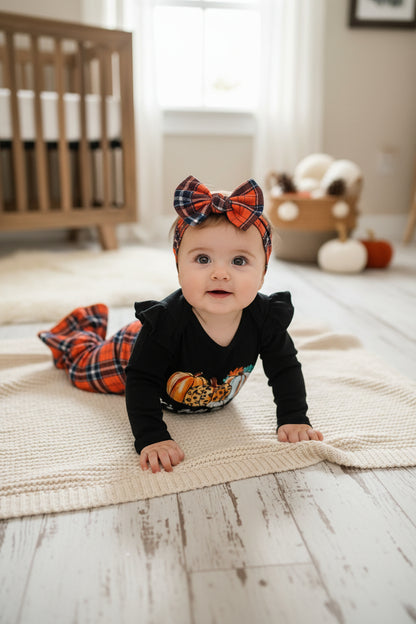 A baby girl's fall-themed two-piece outfit set laid flat on a gray wood plank floor. The set includes a black long-sleeve bodysuit with a pumpkin and leopard print graphic and the text 'It's Fall If u like', paired with orange, navy, and white plaid pants and a matching plaid bow headband.