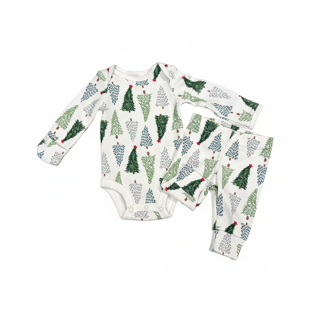 A baby two-piece outfit consisting of a long-sleeve bodysuit and matching pants, featuring an all-over Christmas tree print on a white background, laid flat on a dark wood floor.