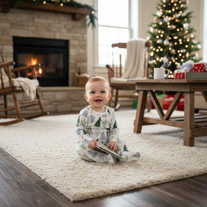 A baby two-piece outfit consisting of a long-sleeve bodysuit and matching pants, featuring an all-over Christmas tree print on a white background, laid flat on a dark wood floor.