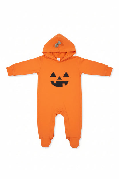 An orange long-sleeve baby bodysuit or pajama set laid flat on a dark wood floor, featuring a black pumpkin face graphic on the chest and a matching hood with a gray zipper.